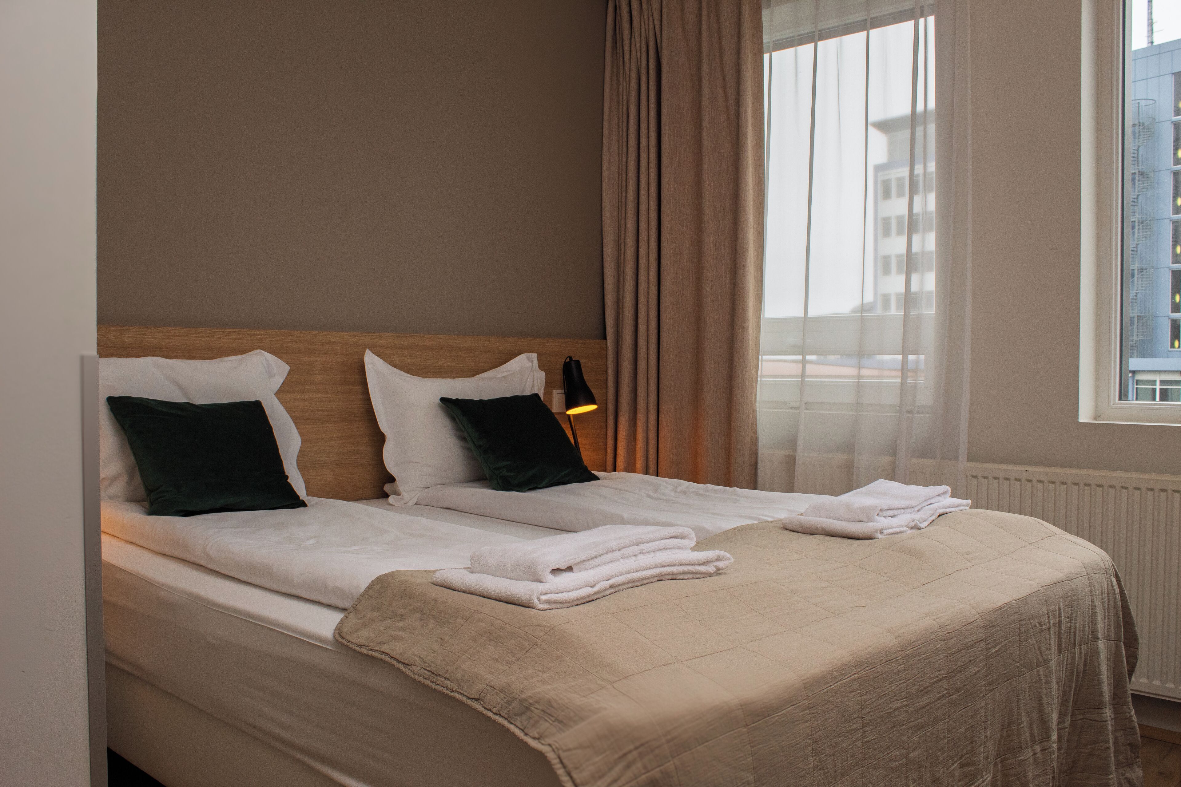 A hotel room with a bed, two pillows, a blanket, and a window with curtains.