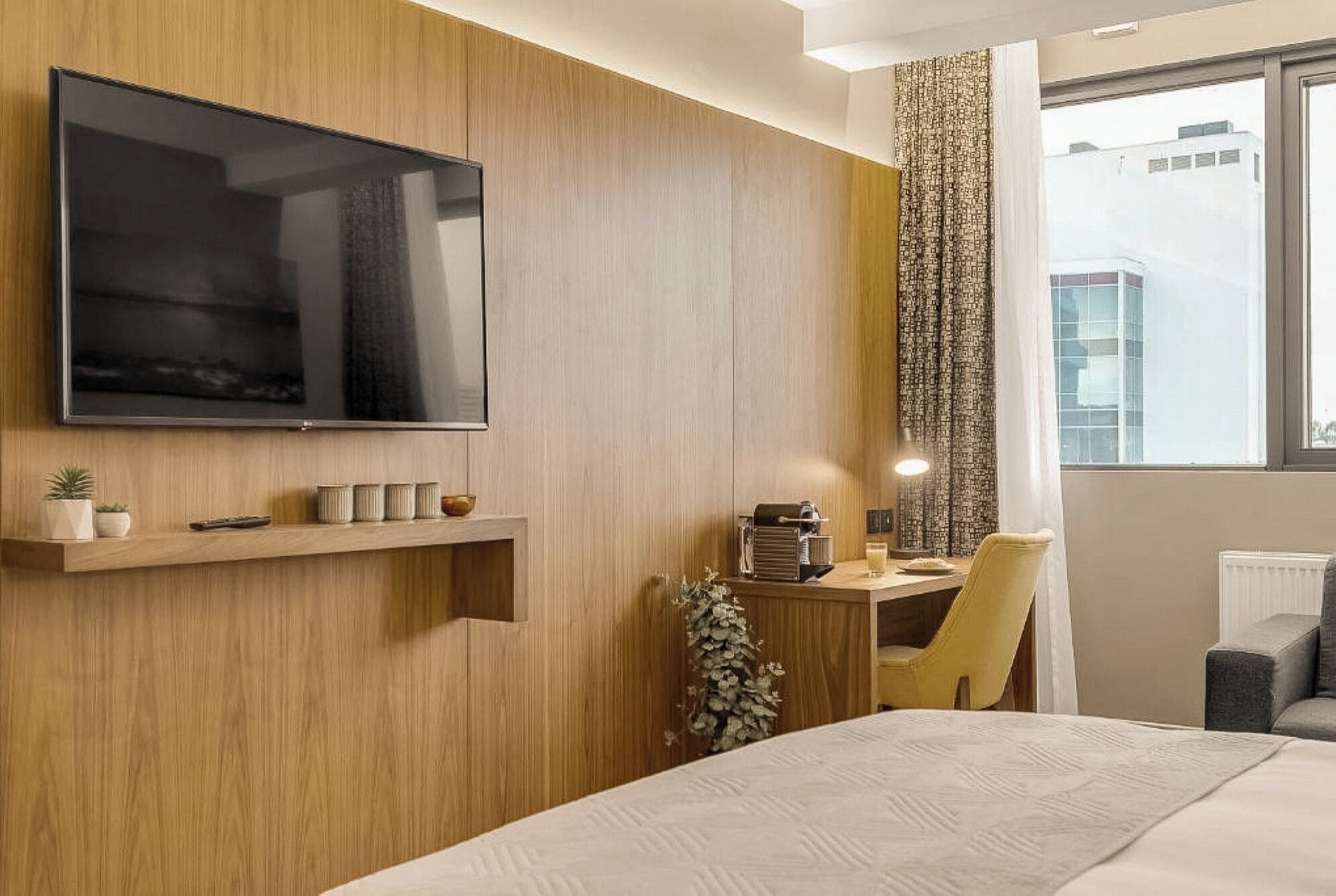 A hotel room with a mounted TV, a small shelf with decorative items, a desk with a lamp, and a chair.
