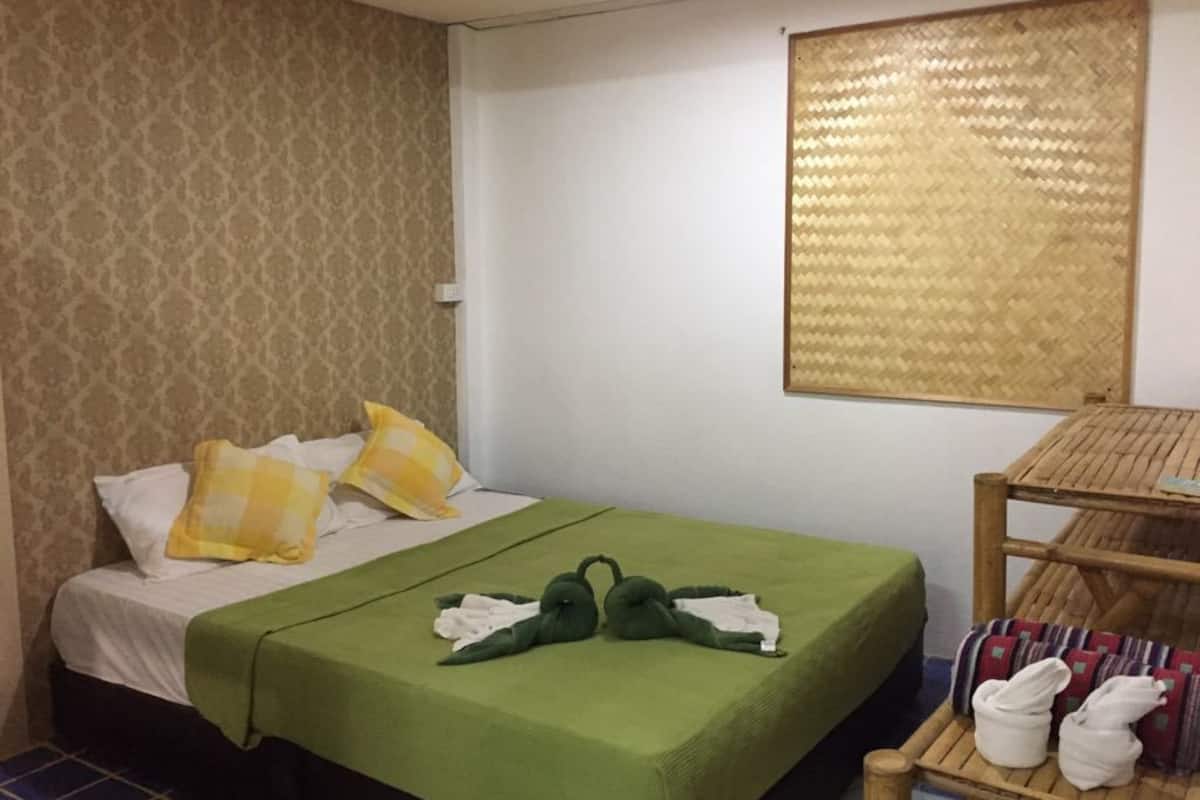 Habitación de hotel con cama, colcha verde, almohadas estampadas, cabecero tejido y una pequeña mesa de madera con toallas.
