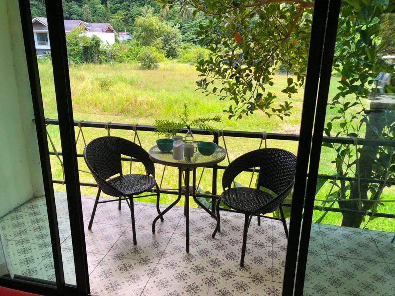 A balcony with two wicker chairs and a small table, overlooking a green landscape with trees and a building in the background.