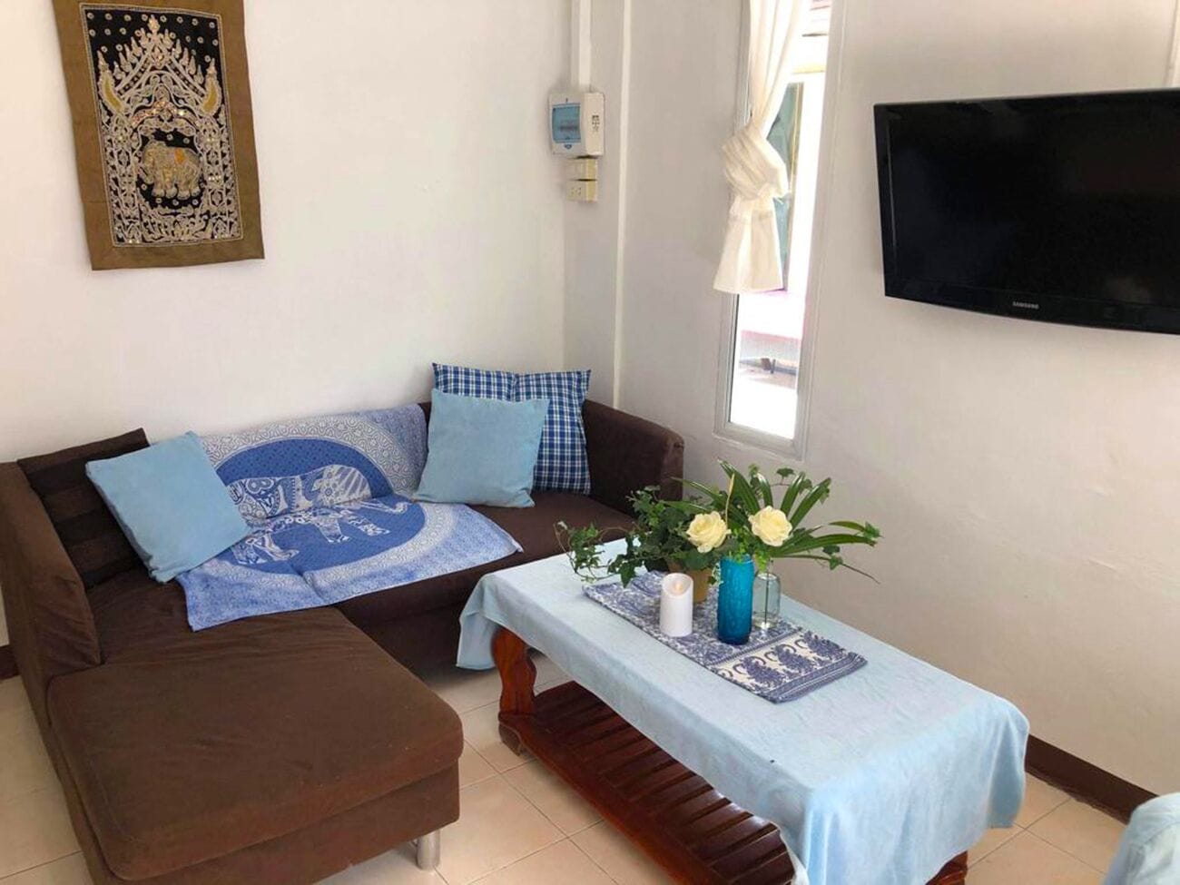 A living room with a brown sofa, blue and white patterned cushions, a small table with a vase of flowers, and a flat-screen TV mounted on the wall.