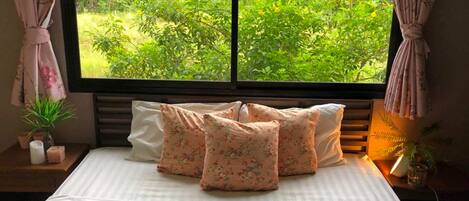 Una cama con almohadas florales, una planta en la mesita de noche y vista a un paisaje verde y exuberante a través de una ventana.
