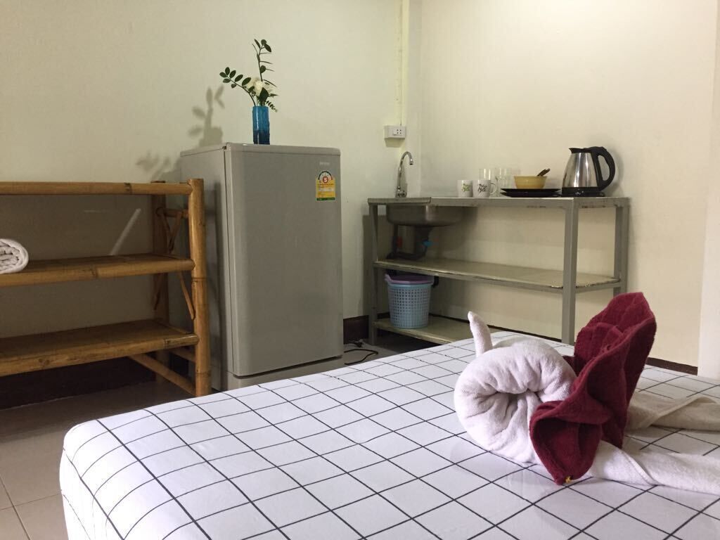 A room with a bed, a small kitchenette with a refrigerator, a sink, and a shelf with kitchenware.