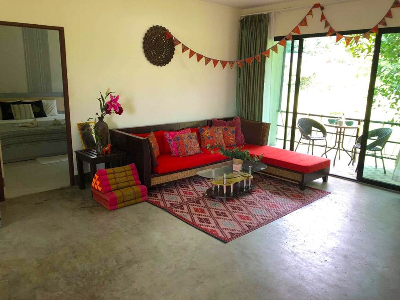 A living room with a red sofa, a glass coffee table, and a patterned rug. There is a large window with curtains and a view of greenery outside.