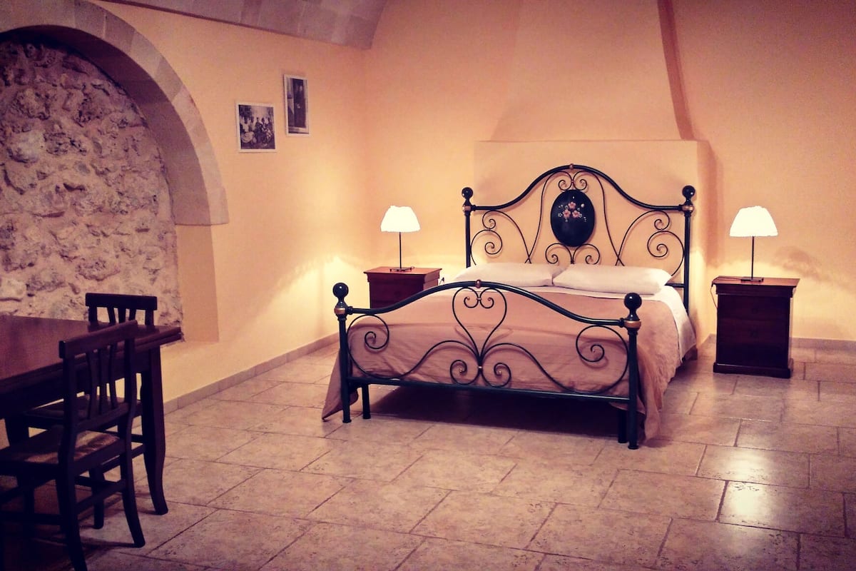 A bedroom with a bed, two bedside tables with lamps, a chair, and a stone wall.