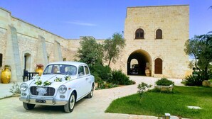 A white vintage car parked in front of a stone building with arched doorways and a tower.