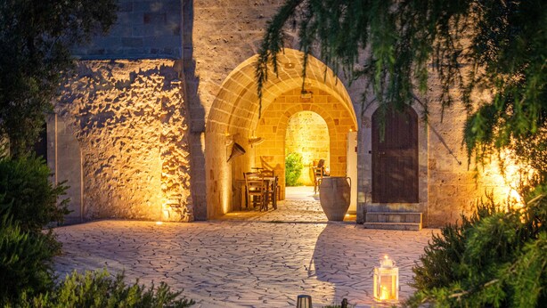An outdoor stone pathway leading to an arched entrance with a table and chairs, surrounded by greenery and lit by lanterns.