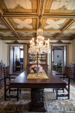Ein Esszimmer mit großem Holztisch, einer verzierten Chandelüster und einem gemusterten Teppich.