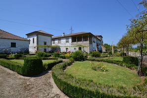 Um edifício tradicional de dois andares com telhado de azulejos, rodeado por um jardim bem cuidado com sebes e canteiros de flores.