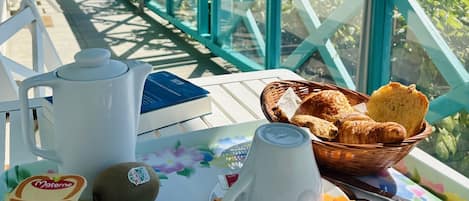 A table with breakfast items, a white mug, a basket of bread, and a cup of coffee on a balcony with a turquoise railing and a view of greenery and buildings.