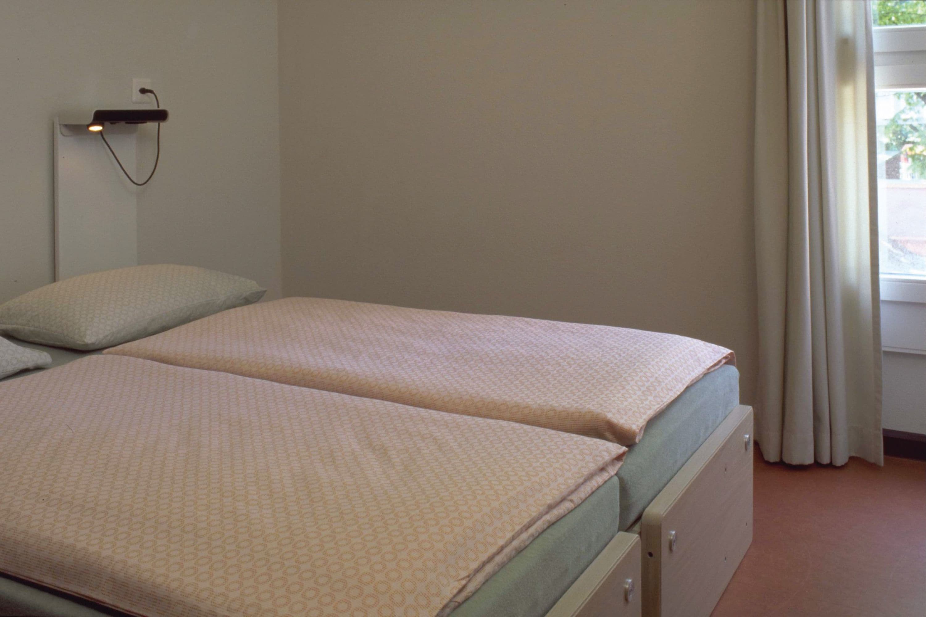 Una habitación sencilla con una cama individual, pared lisa, lámpara de noche y ventana con cortinas.