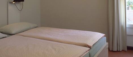 A simple, single-bed room with a plain wall, a bedside lamp, and a window with curtains.