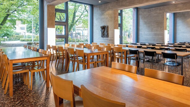 A spacious indoor area with multiple wooden tables and chairs, large windows offering views of trees and buildings outside. - Youth Hostel Zurich (Zürich)