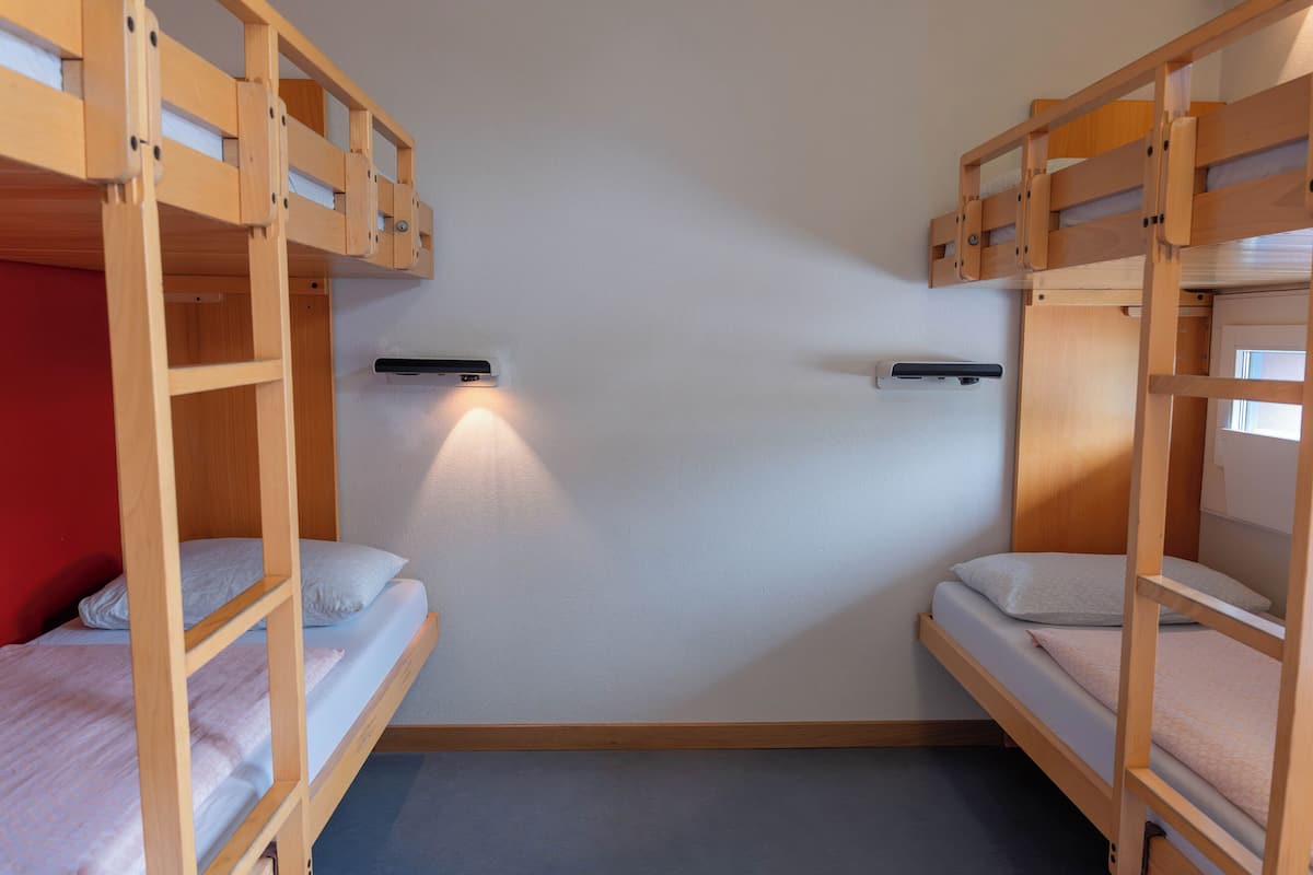 Una habitación con dos literas, cada una con una almohada y un pequeño estante. Hay una lámpara de pared entre las camas.