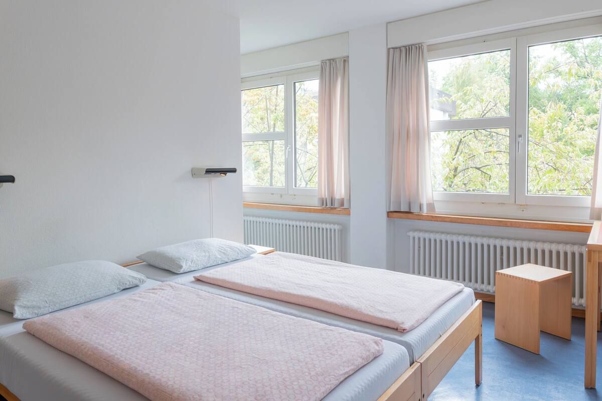 Una habitación con dos camas, una mesita de madera, un radiador y ventanas grandes con cortinas.