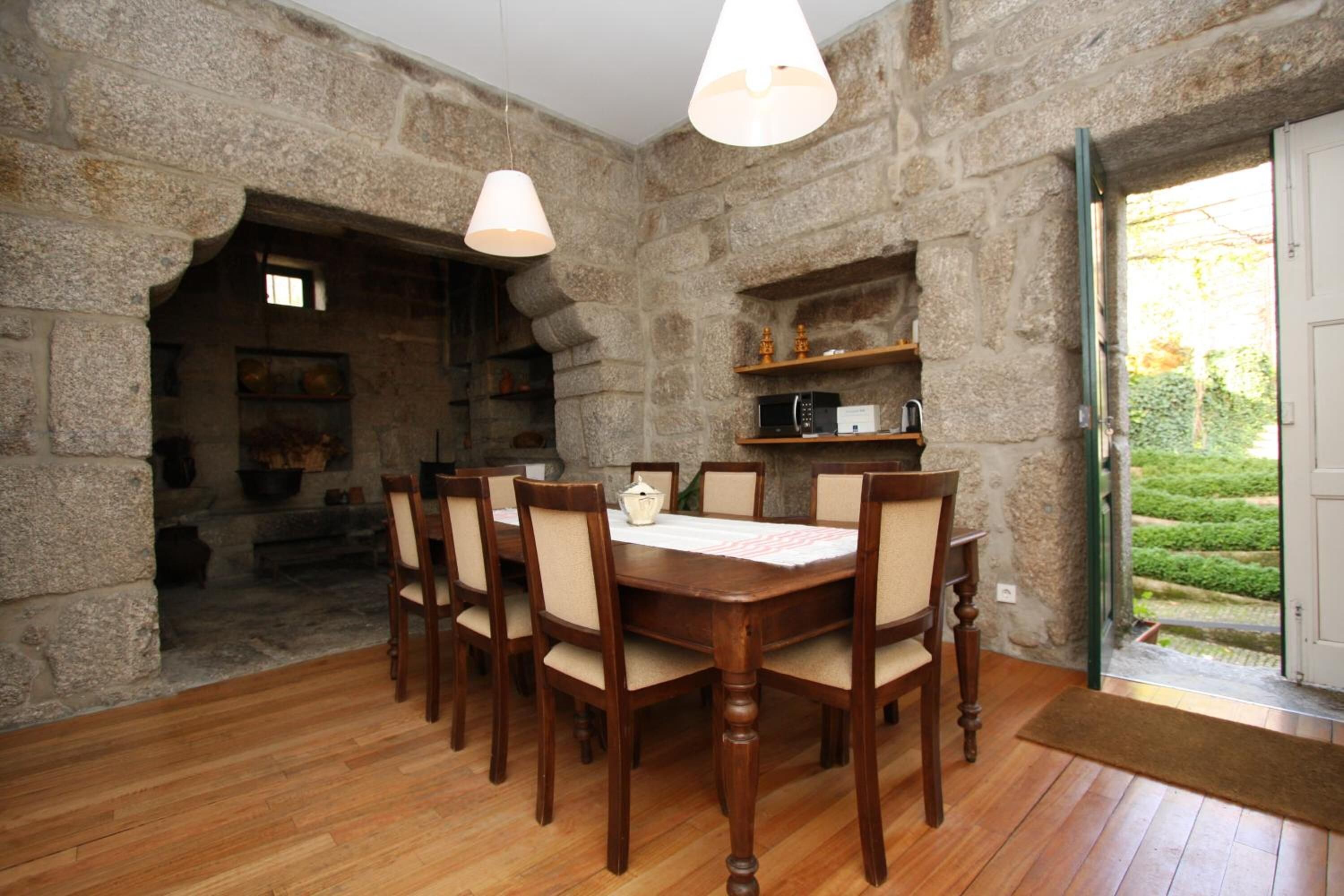 A rustic dining room with a stone wall, wooden dining table, and chairs. There is a doorway leading to a garden.