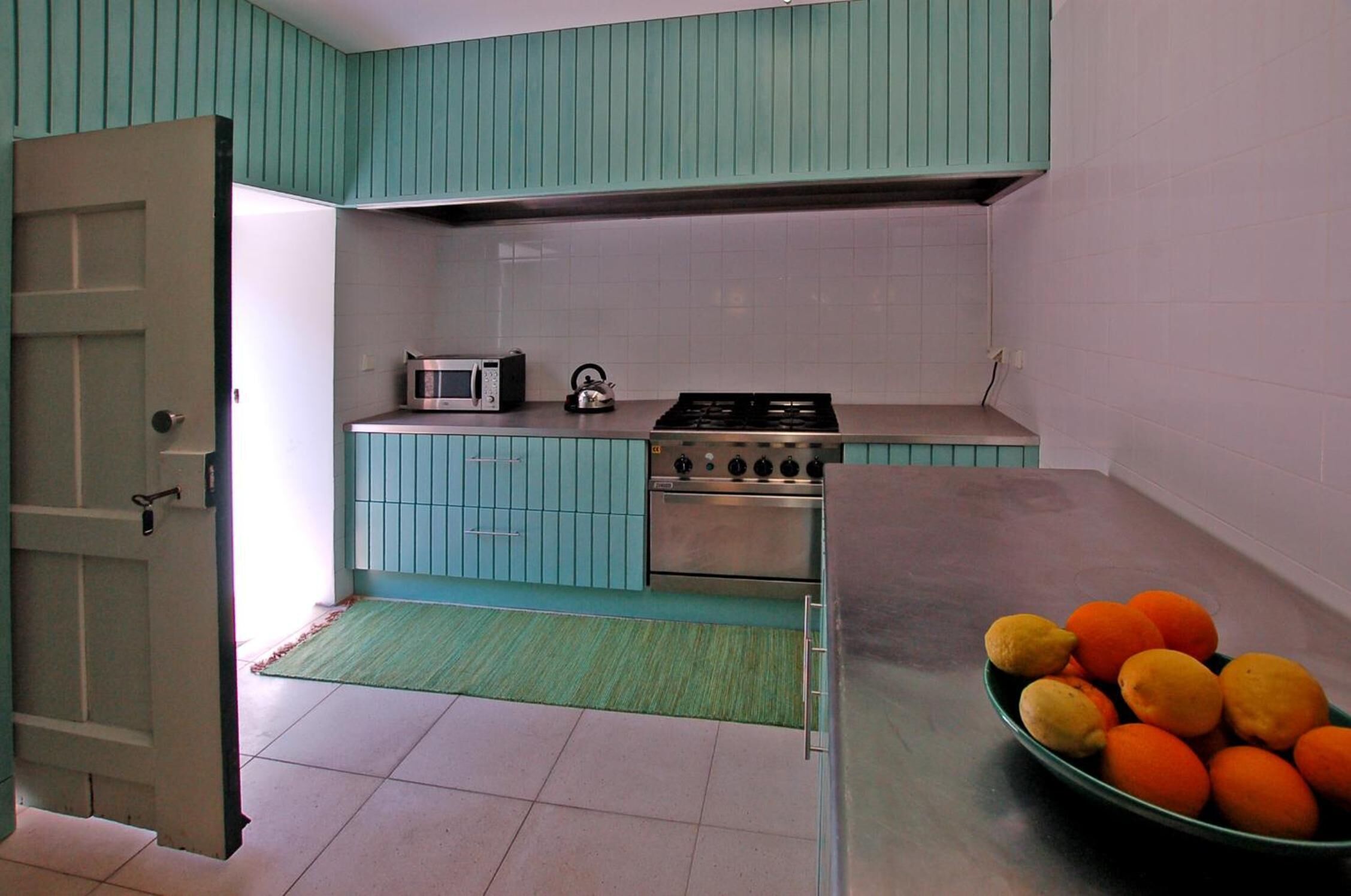 A kitchen with teal cabinets, a microwave, a kettle, and a stove. There is a bowl of oranges on the counter.
