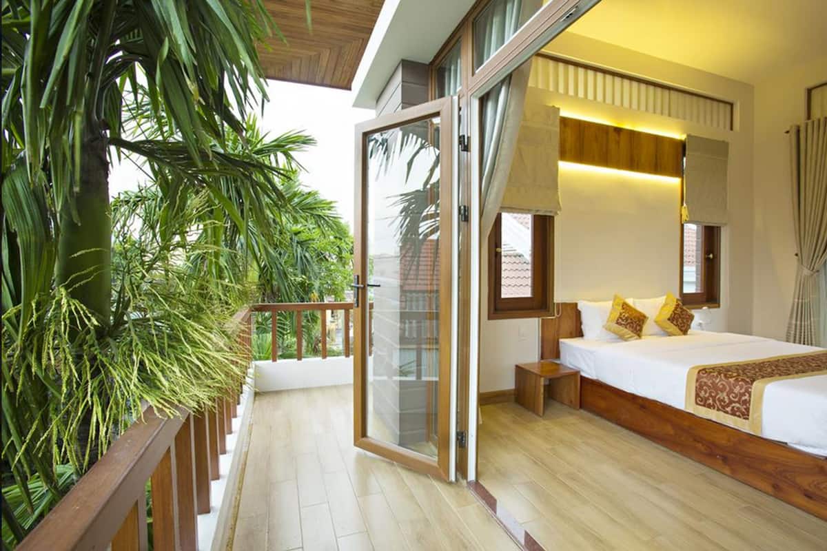 A bedroom with a large bed, a wooden floor, a sliding glass door leading to a balcony with plants, and a view of the outside.