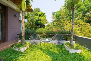 A patio with three metal chairs and a glass table, surrounded by lush greenery and a wooden fence. - Hanging Garden Hoi An (Da Nang)