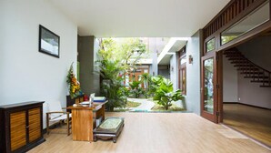 A spacious indoor area with a wooden floor, a reception desk, a chair, and a small table. There is a garden view through a glass door. - Hanging Garden Hoi An (Da Nang)