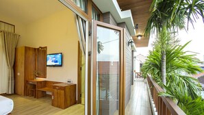 A modern room with a wooden desk, a television, and a sliding glass door leading to a balcony with plants. - Hanging Garden Hoi An (Da Nang)