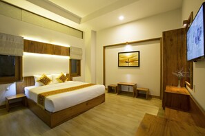 A modern hotel room with a large bed, wooden headboard, bedside tables, a small desk, and a TV mounted on the wall. - Hanging Garden Hoi An (Da Nang)