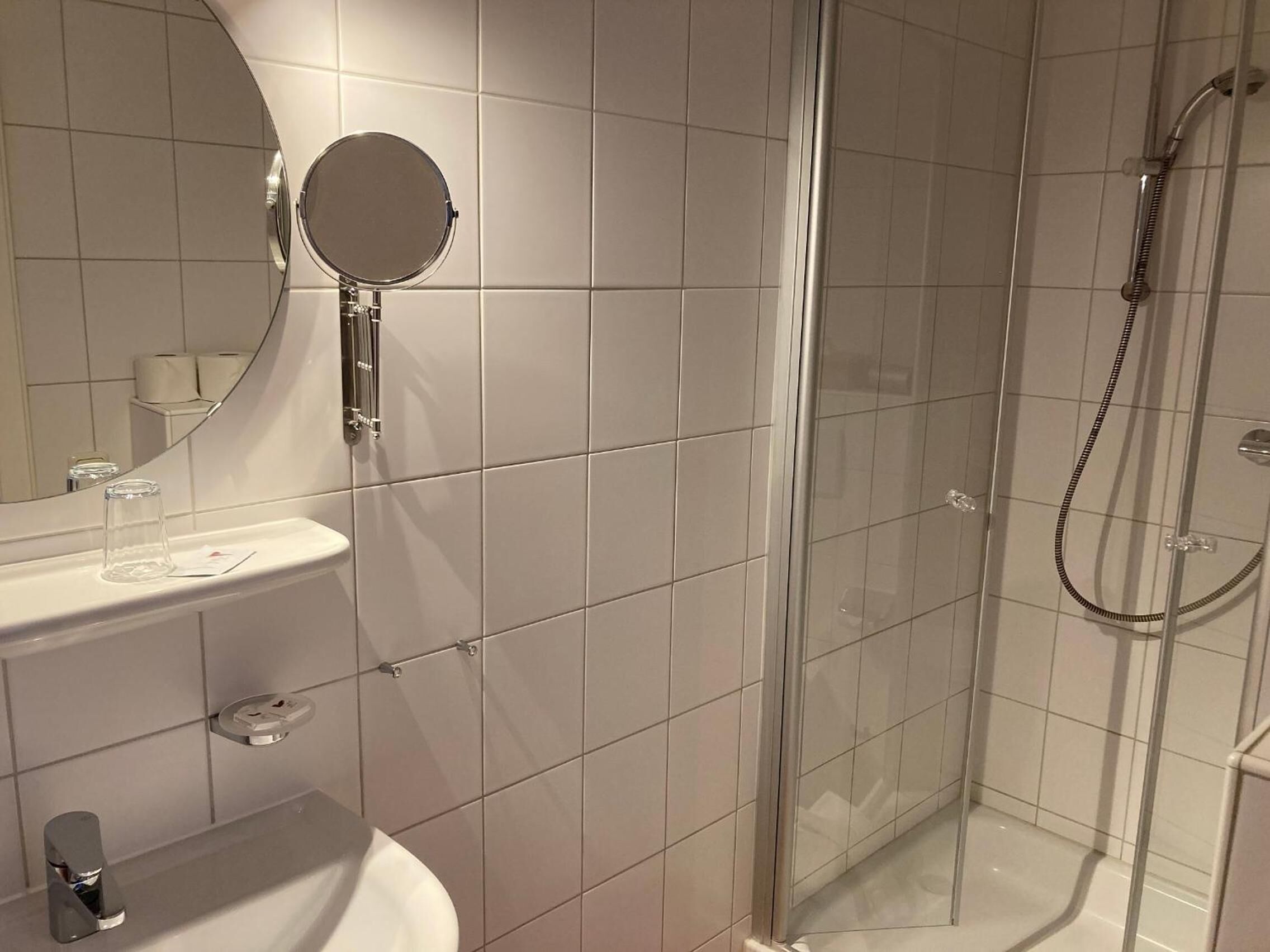 A modern bathroom with a glass-enclosed shower, a round mirror, and a white sink with a soap dispenser.