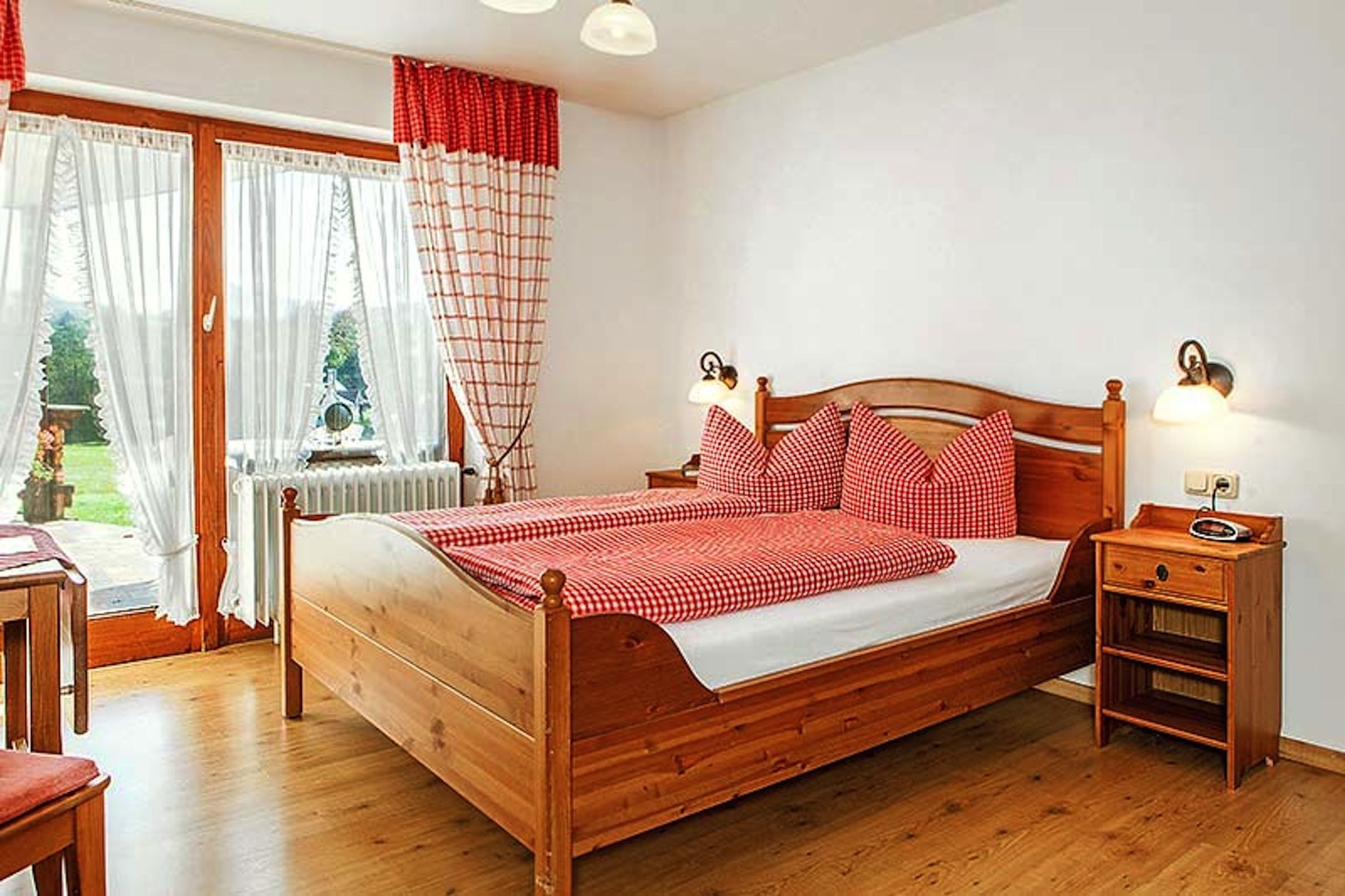 A wooden bed with checkered bedding, a bedside table with a lamp, and a view of a garden through a window with curtains.