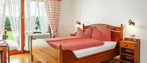 A wooden bed with checkered bedding, a bedside table with a lamp, and a view of a garden through a window with curtains.