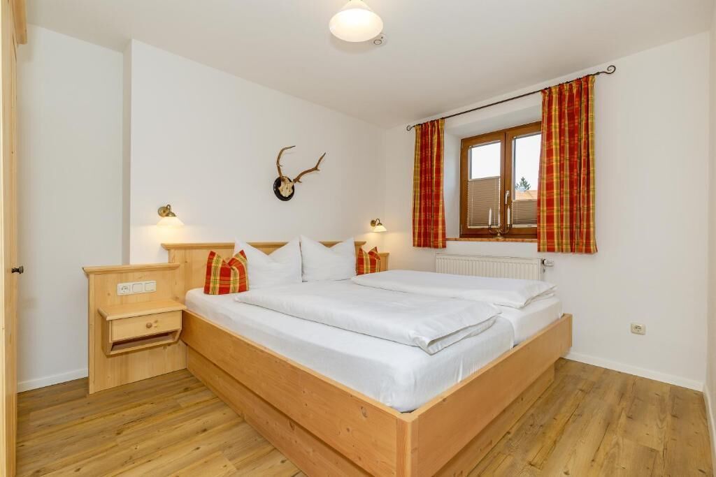 A bedroom with a wooden bed, a nightstand, a wall-mounted deer head decoration, and a window with red curtains.