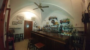 A bar area with a wooden counter, stools, a ceiling fan, and shelves with bottles. - Hostal Marqués de Zahara (Zahara de la Sierra)