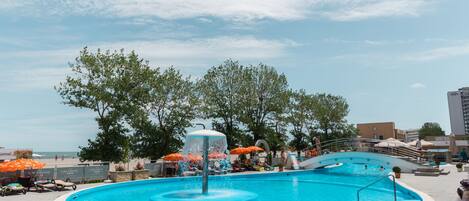 A swimming pool with a water fountain, surrounded by lounge chairs and umbrellas, with trees and buildings in the background.