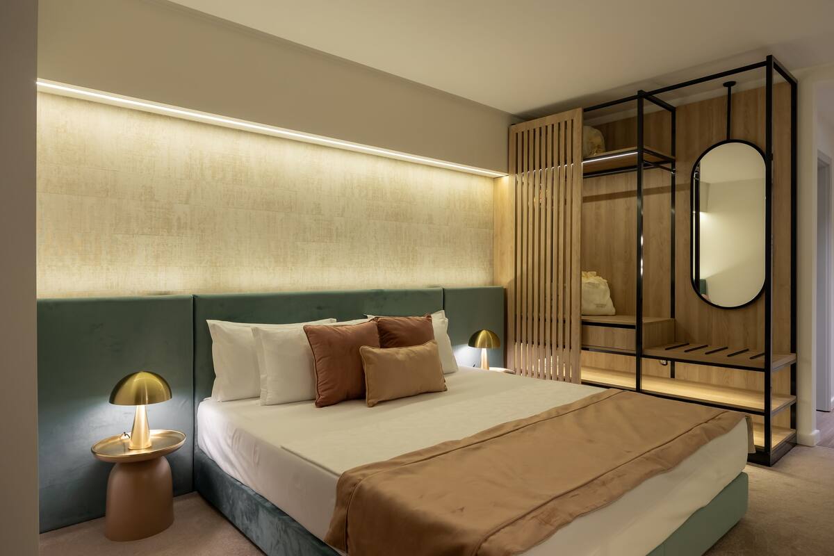 Moderni hotellihuone, jossa on suuri sänky, yöpöydällä olevat lamput, peilillä varustettu vaatekaappi ja sängynpääty, jossa on sisäänrakennettu valaistus.