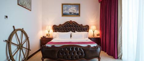 A large bed with a dark headboard, two bedside tables with lamps, a large wooden wheel, and a painting of a ship on the wall.