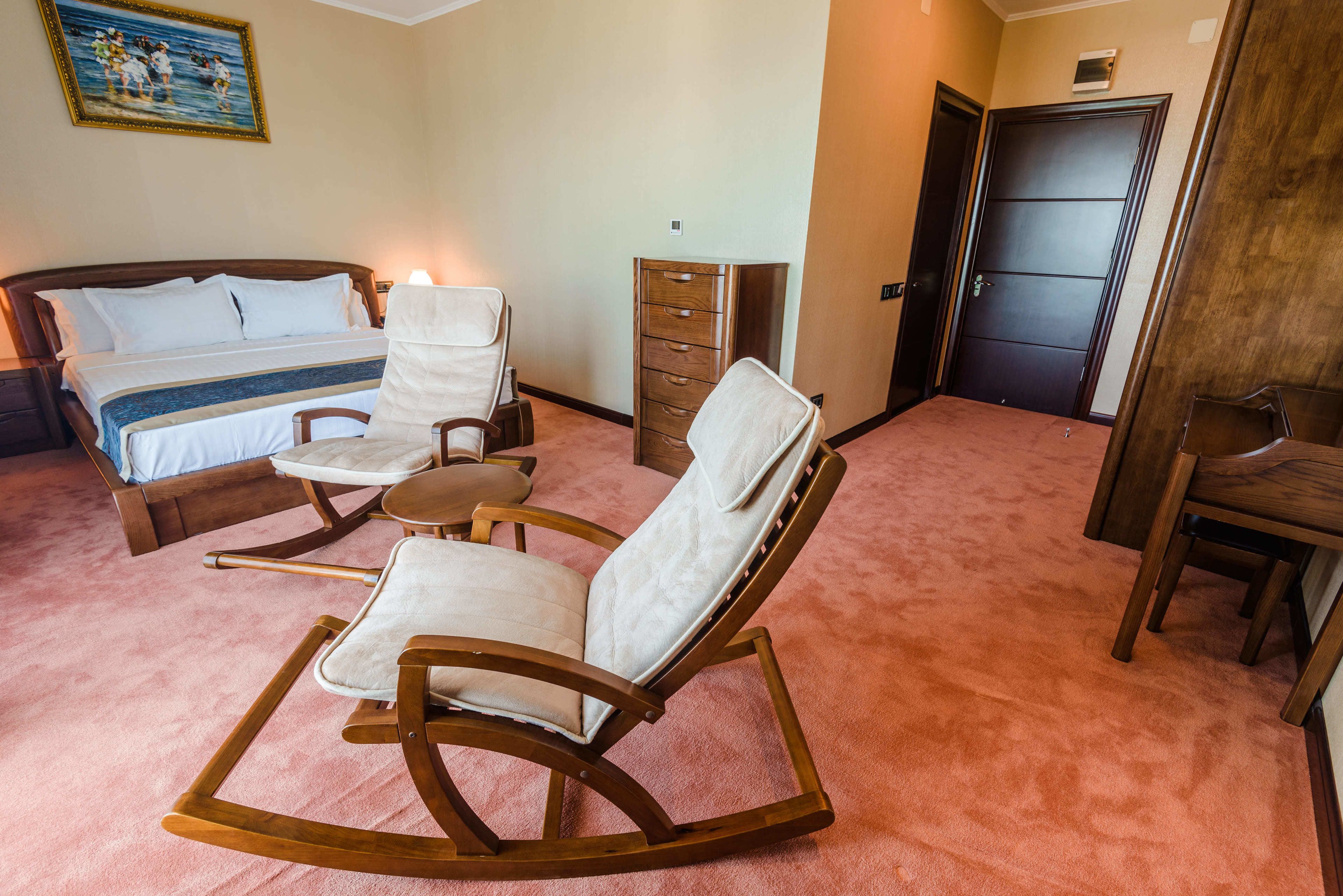 A bedroom with a bed, a rocking chair, a small table, a dresser, and a door.