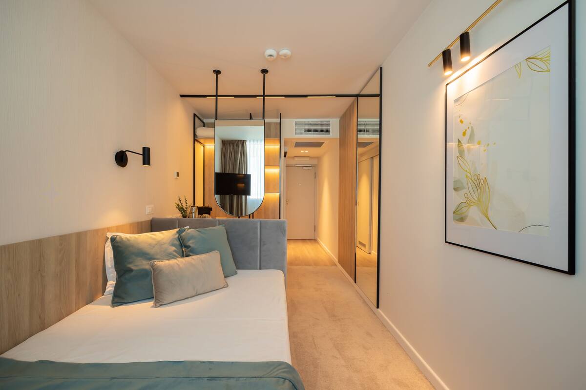 Moderni hotellihuone, jossa on sänky, taulutelevisio, suuri kehystetty taideteos ja käytävä, joka johtaa kylpyhuoneeseen.