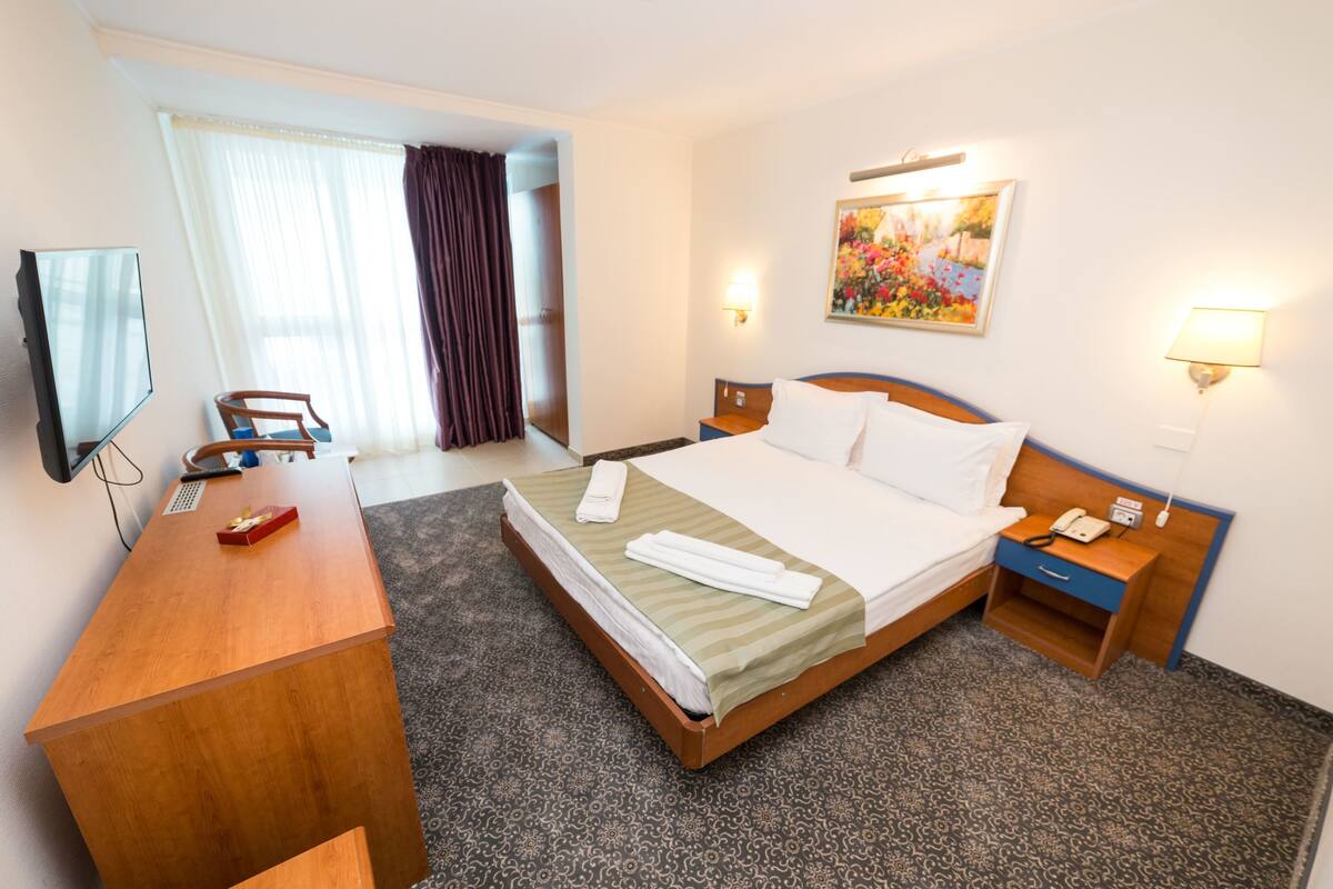 Hotellihuone, jossa on sänky, työpöytä, tuoli, televisio, taulu ja ikkuna, jossa on verhot.