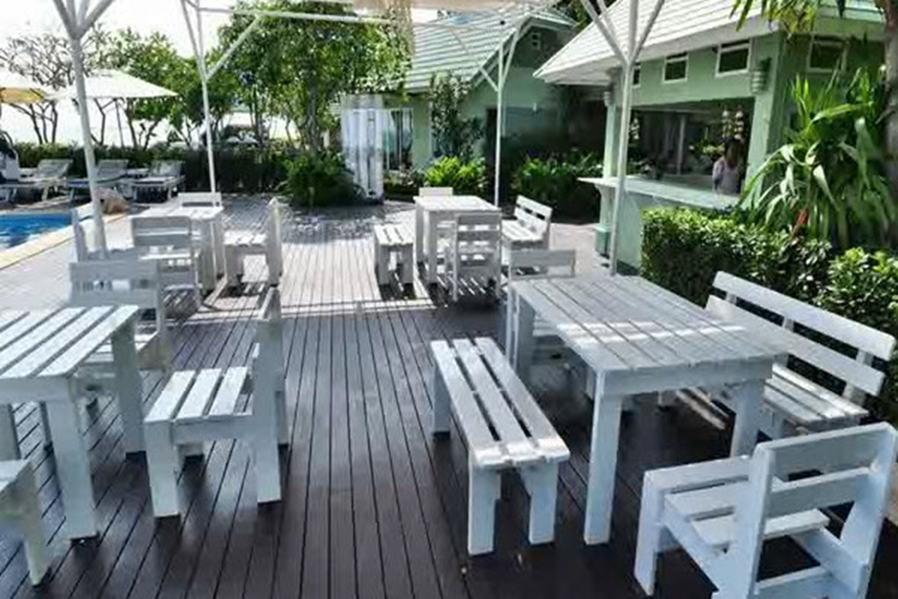 Una terraza de madera con mesas y sillas blancas, una piscina y un edificio con techo verde.