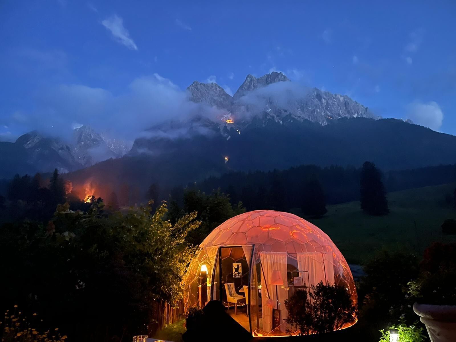 A dome-shaped tent with a glass door, surrounded by trees and a mountain backdrop.
