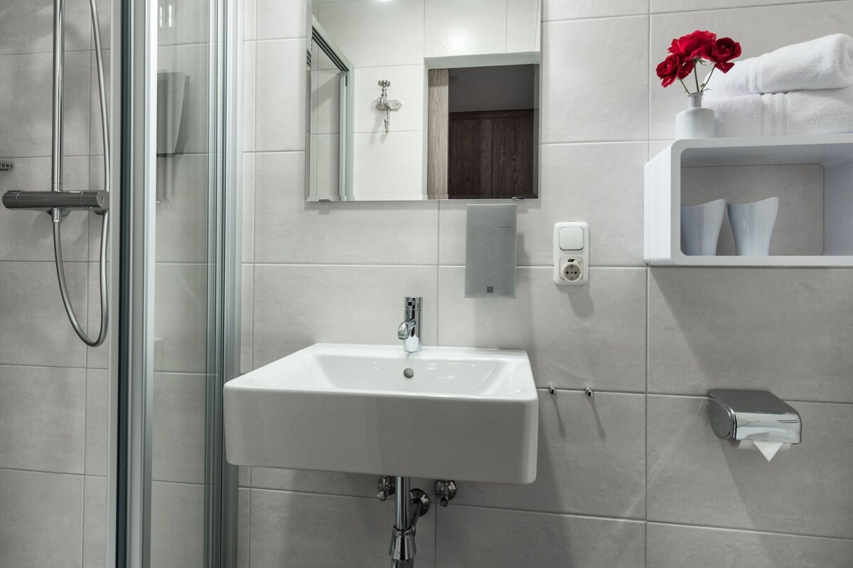 Un baño moderno con cabina de ducha de vidrio, un lavamanos blanco y un perchero con un jarrón de flores rojas.
