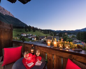 A balcony with a table set for two, featuring a bottle of wine, glasses, playing cards, and candles. The view includes a village with houses and mountains in the background. - Bayern Resort Hotel (Grainau)