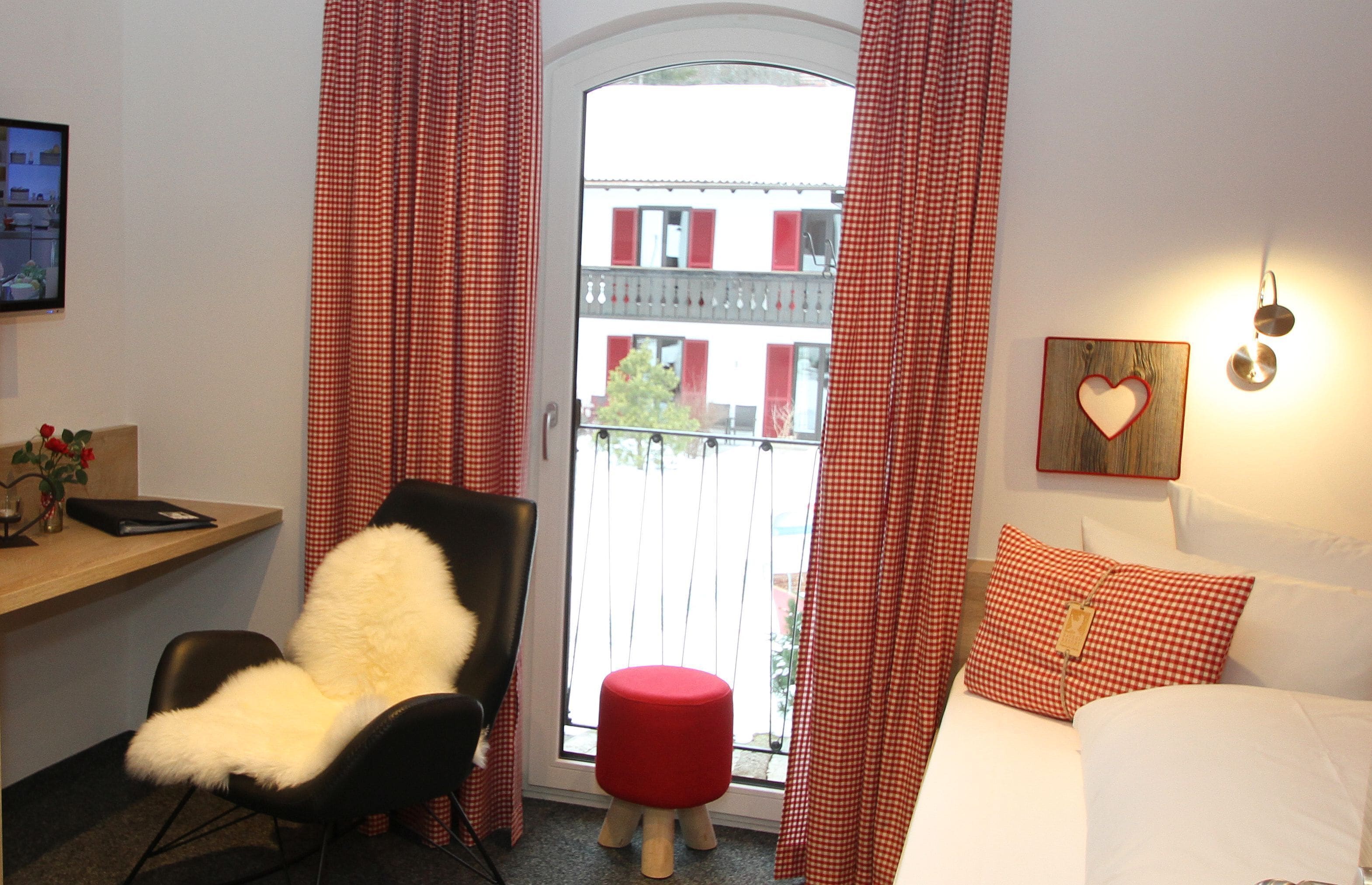 Una habitación de hotel con una silla negra, un reposapiés rojo, una cama con ropa de cama blanca, una decoración con forma de corazón en la pared y una ventana con cortinas a cuadros rojos.