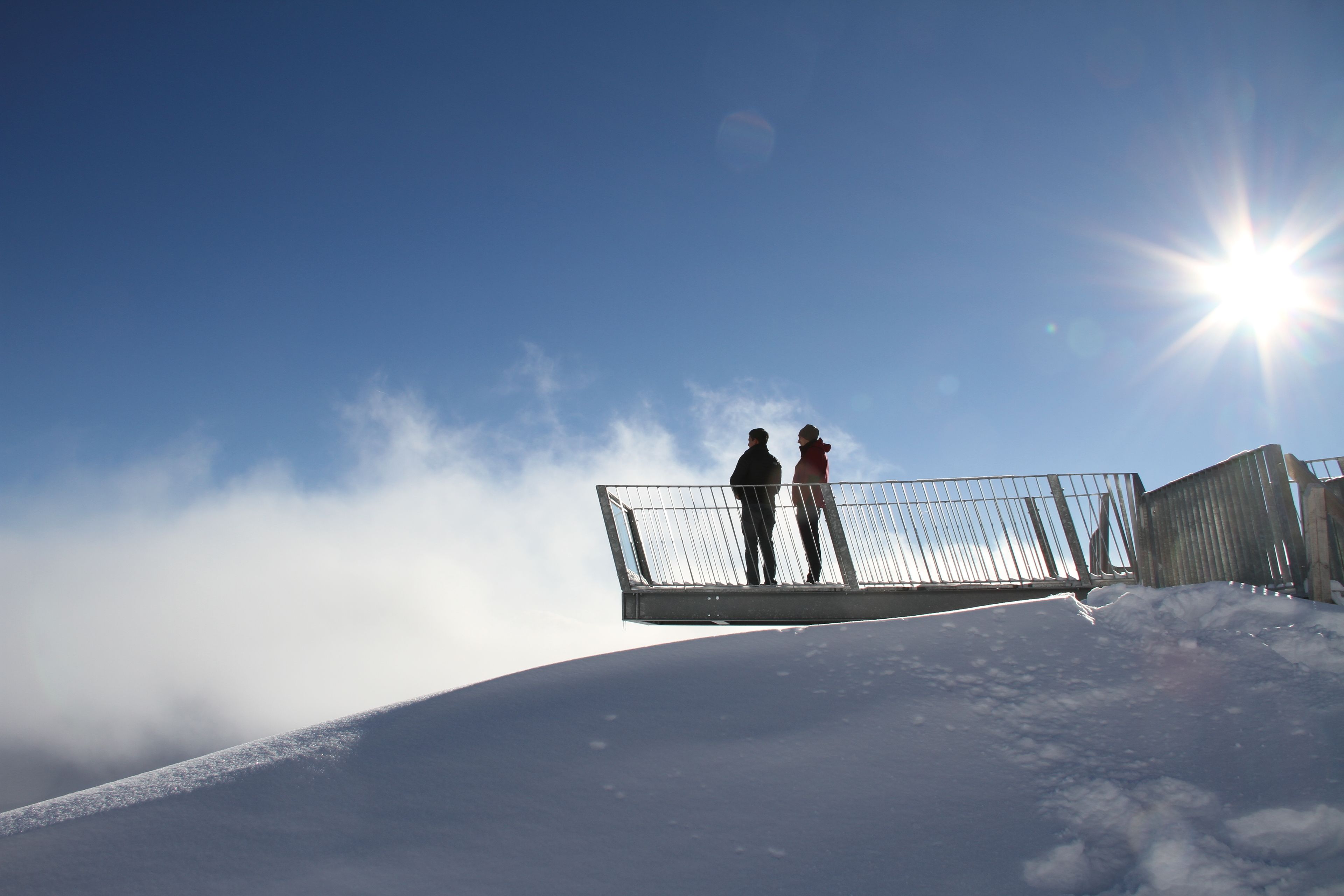 Dos personas de pie en una ladera cubierta de nieve, con una valla metálica, bajo un cielo despejado con el sol brillando.