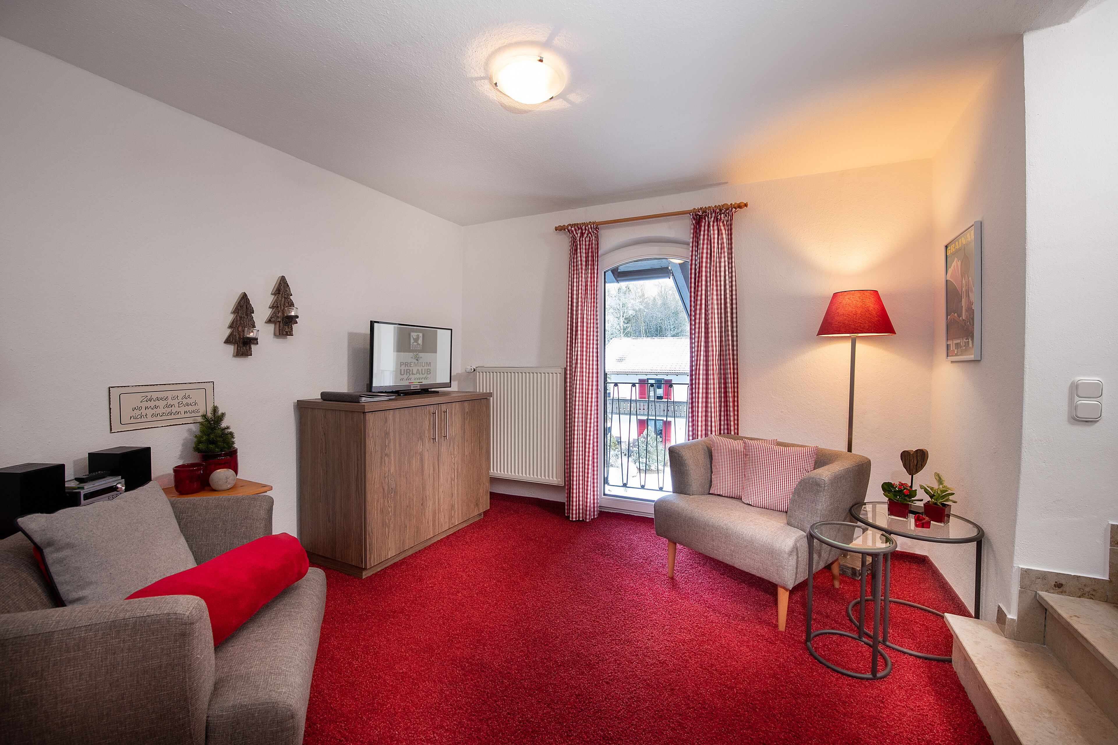 A living room with a red carpet, a grey sofa, a small table with a lamp, and a window with red curtains.