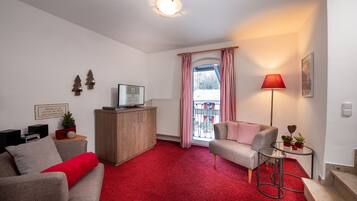 A living room with a red carpet, a grey sofa, a small table with a lamp, and a window with red curtains.