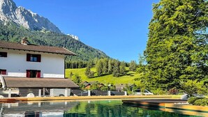 Una piscina con juguetes inflables, montañas al fondo y un edificio de fachada blanca.