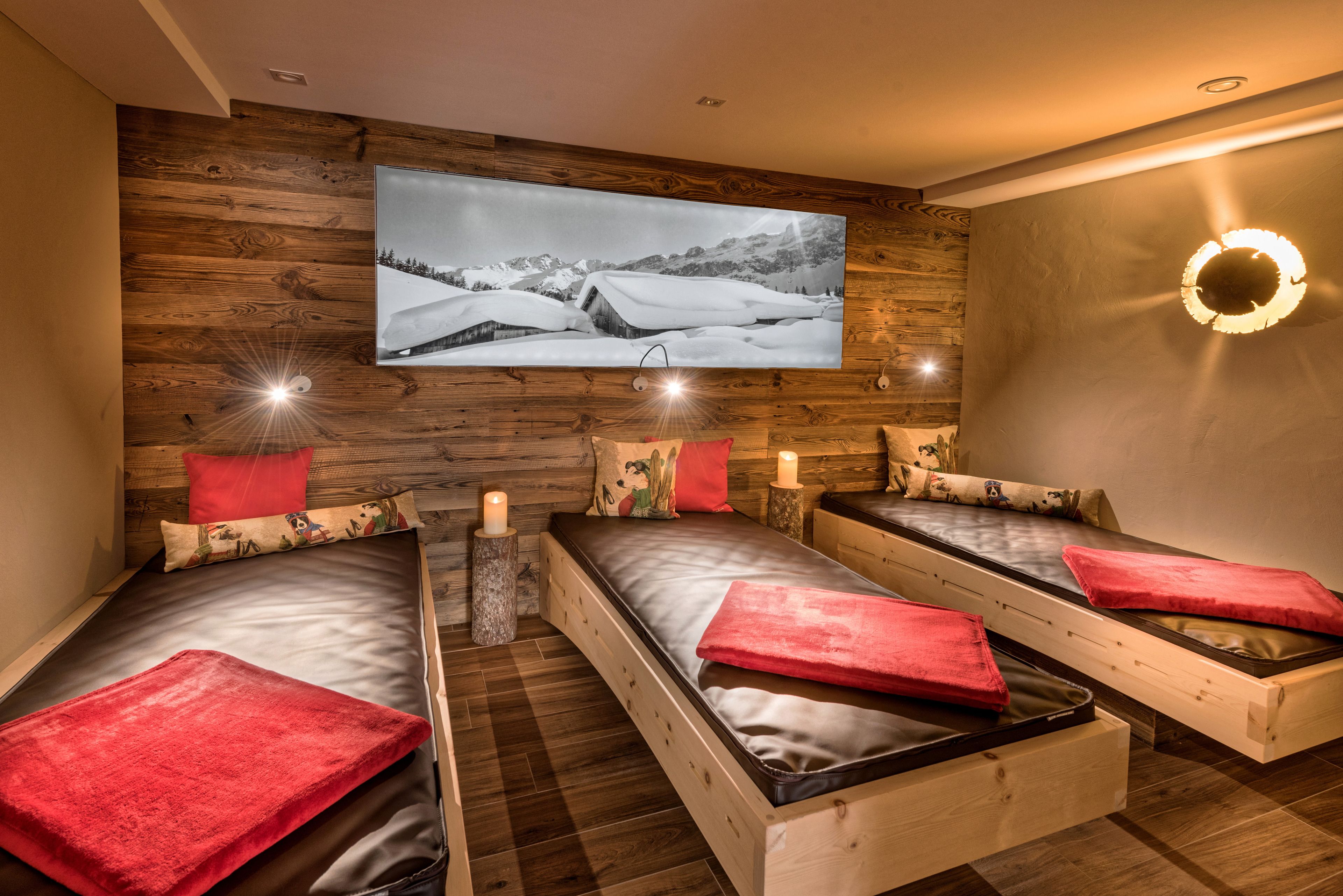 Quarto com três camas de madeira, cada uma com travesseiro vermelho e uma pequena mesa de cabeceira com uma vela. Uma televisão grande fixada na parede exibe uma paisagem nevada.