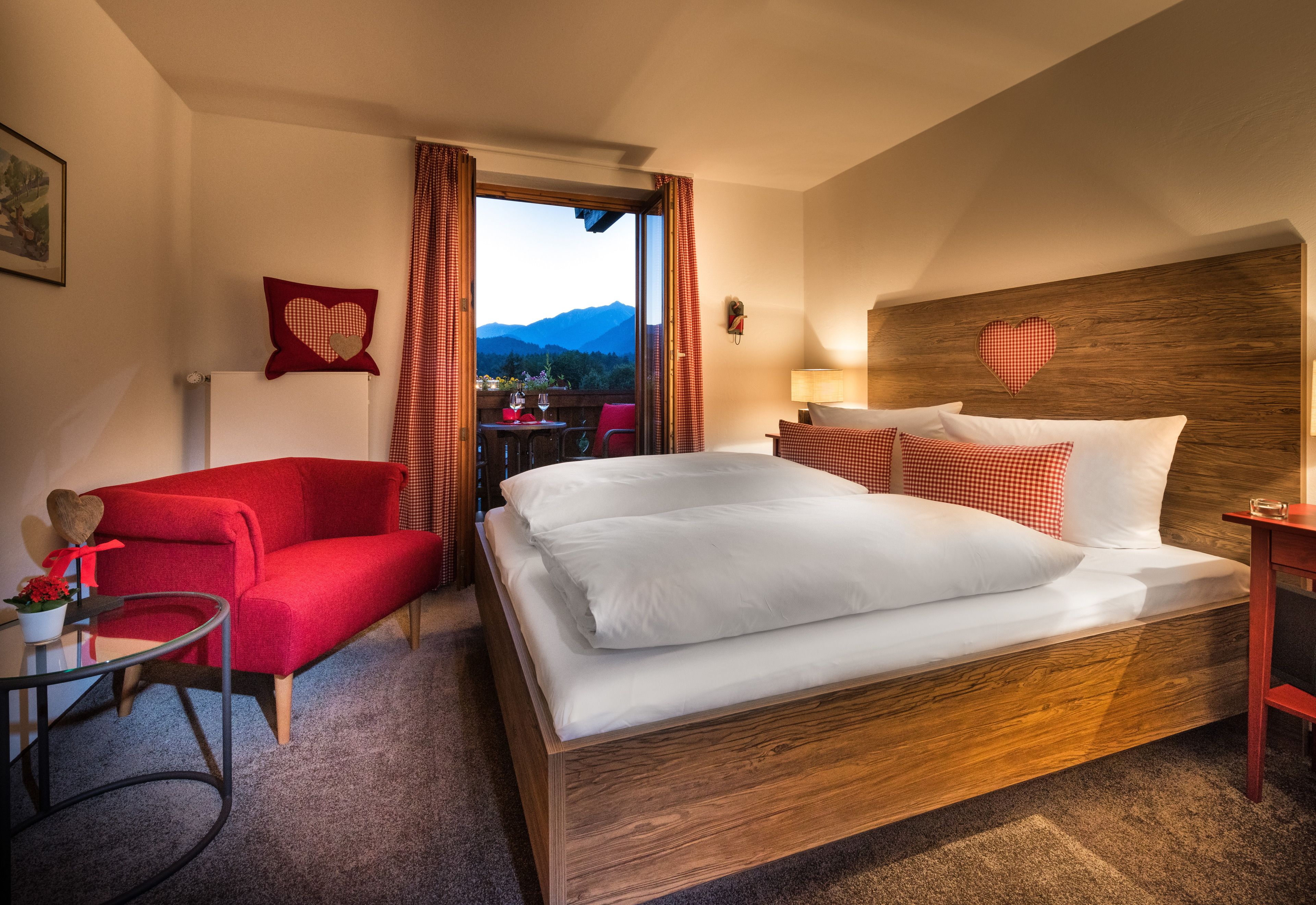 Une chambre d’hôtel avec un grand lit, un fauteuil rouge, une petite table sur laquelle est posé un vase de fleurs, et une vue sur les montagnes par la fenêtre.