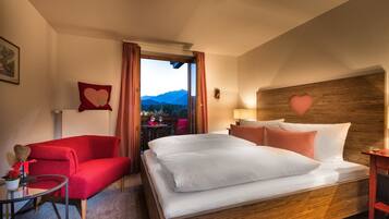 Une chambre d’hôtel avec un grand lit, un fauteuil rouge, une petite table sur laquelle est posé un vase de fleurs, et une vue sur les montagnes par la fenêtre.