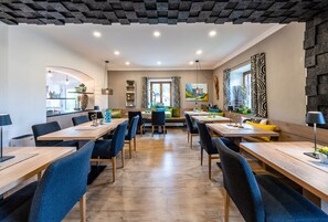 A modern dining area with wooden tables and blue chairs, a kitchen in the background, and a large mirror on the right wall. - Bayern Resort Hotel (Grainau)